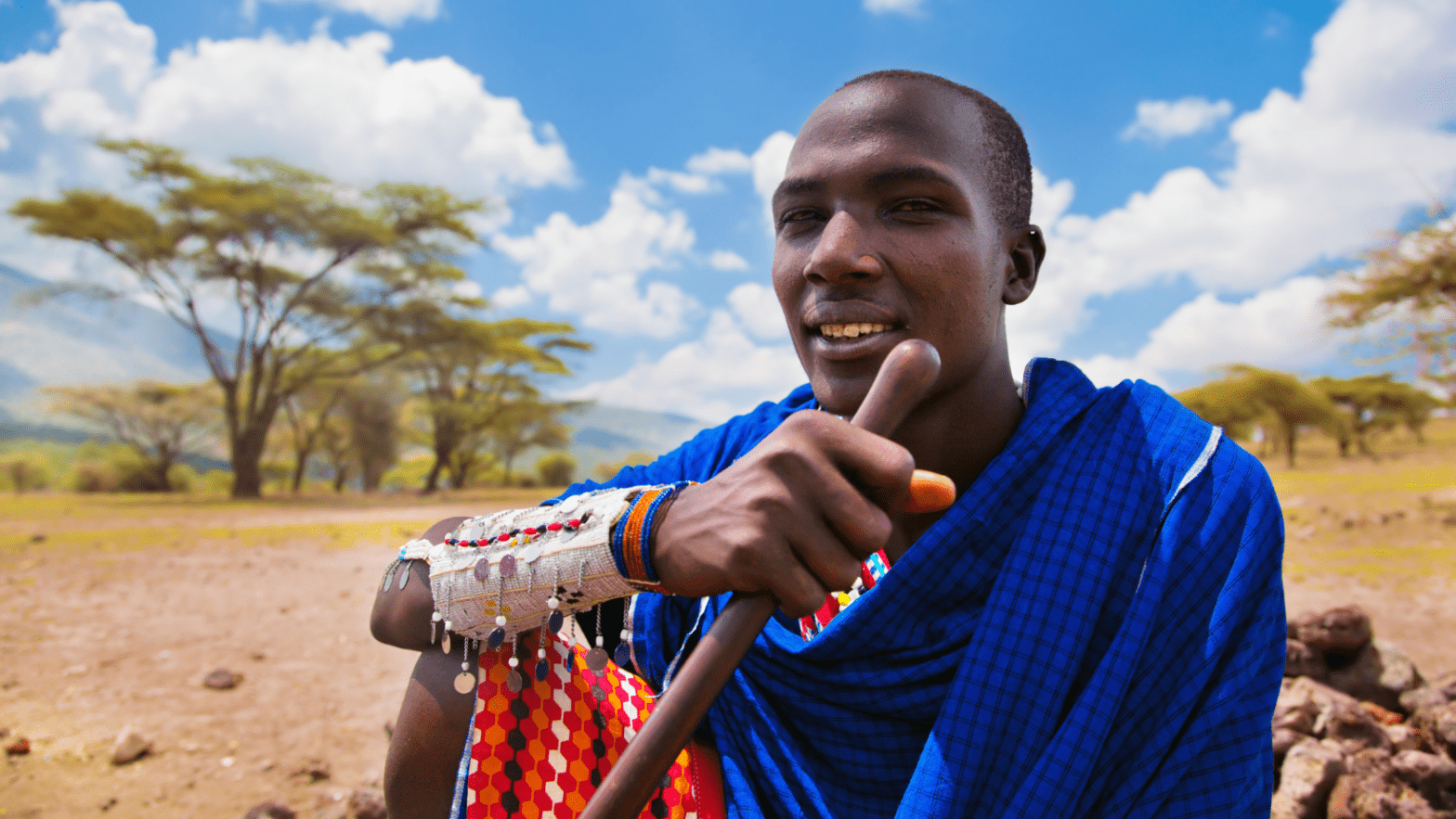 Smiling Tanzanian man in his 30's wearing colorful traditional African outfit sits in Serengeti.