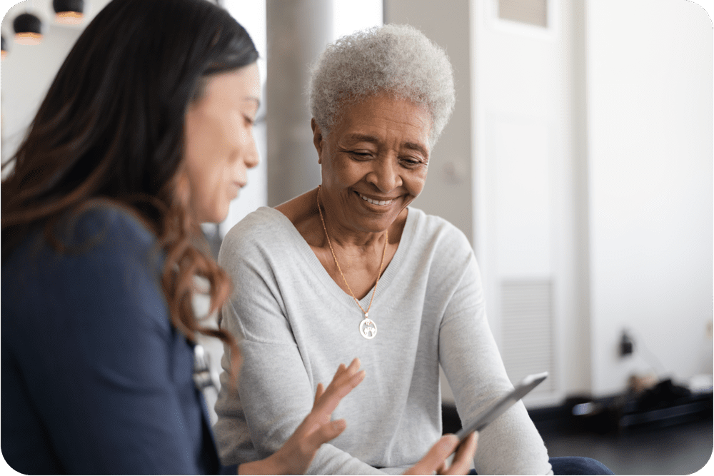 Senior woman in grey sweater smiles as she looks at tablet that medical assistant is holding and referencing to. The Medical assistant helping the patient complete digital registration through AdviNOW's ai assistant digital front door for easy patient intake.